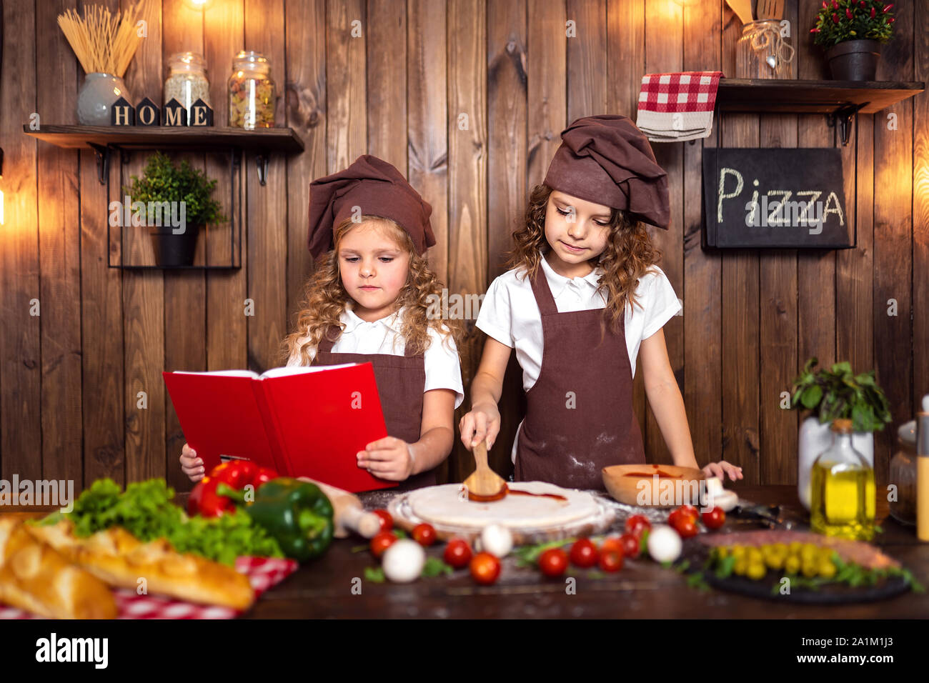 Cute girls in aprons and hats reading recipe book together while ...
