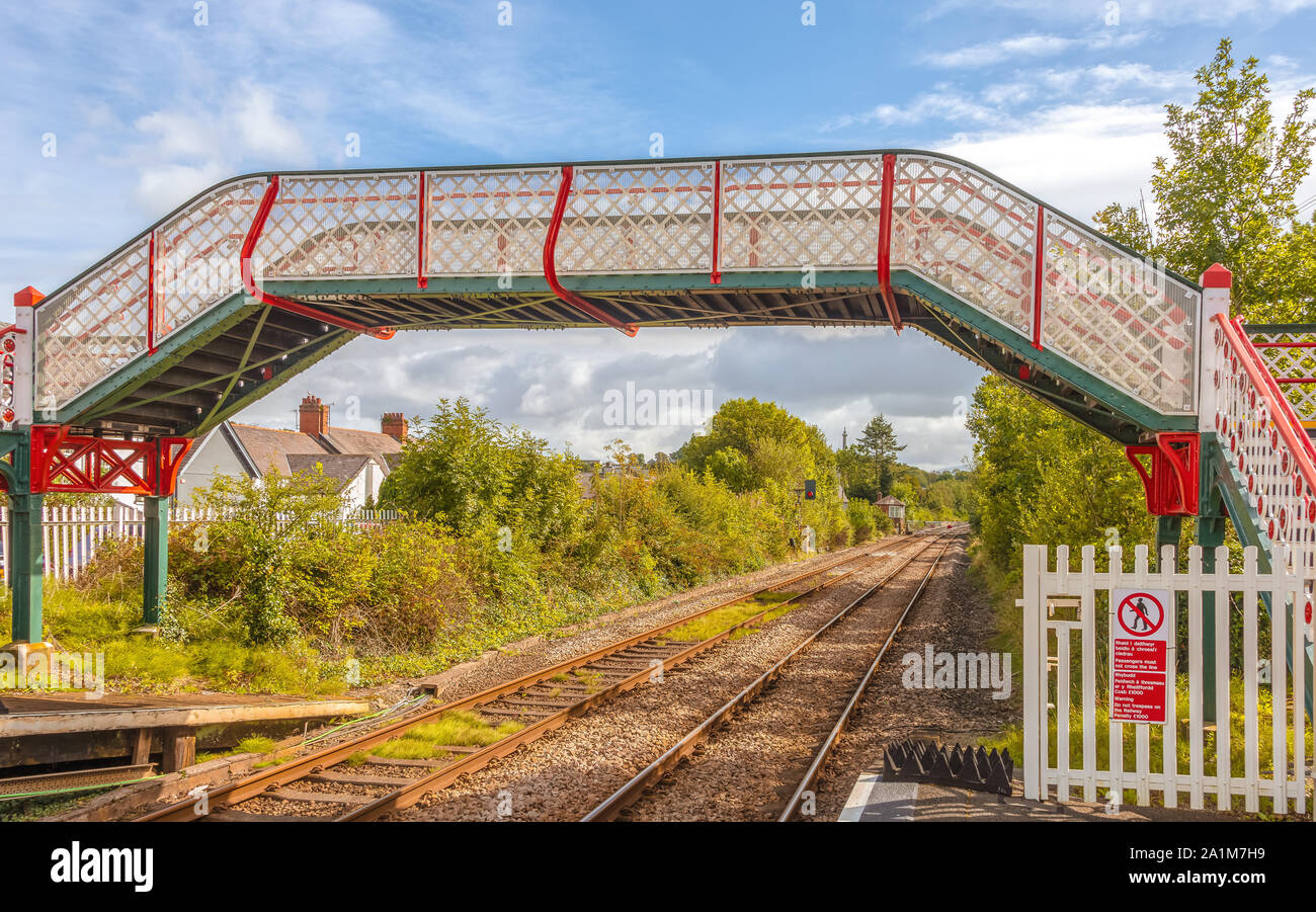 A colourful railway station footbridge at Llanfairpwllgwyngyll, Anglesea in Wales.   Railway lines run underneath and a blue sky with clouds overhead. Stock Photo