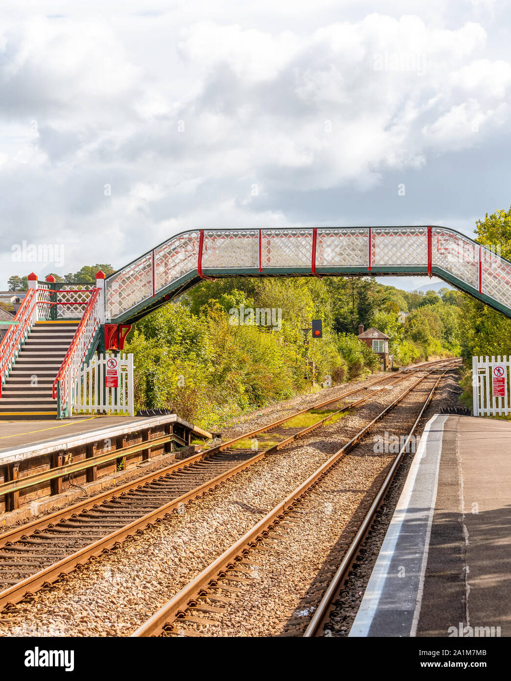 A colourful railway station footbridge at Llanfairpwllgwyngyll, Anglesea in Wales.   Railway lines run underneath and a blue sky with clouds overhead. Stock Photo