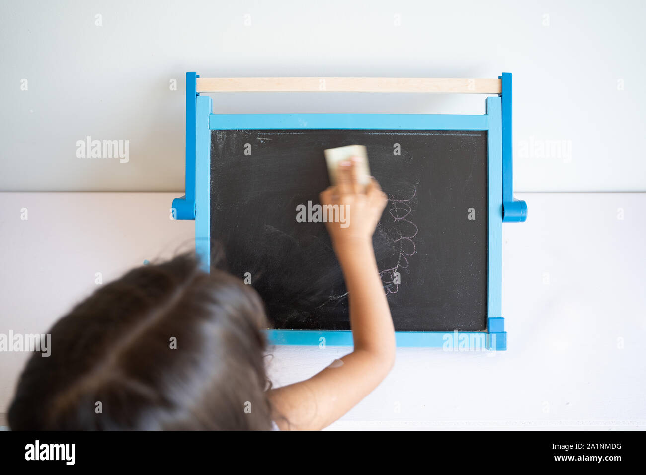 Small girl with long brown hair erasing small blackboard with white surroundings seen from behind Stock Photo
