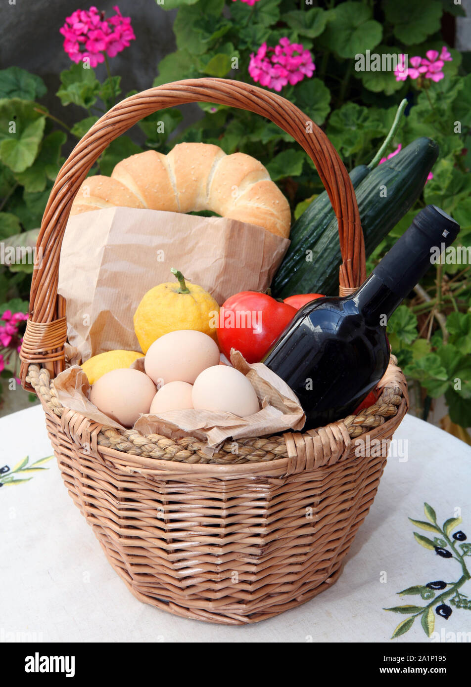 Wicker shopping basket with traditional Greek goods, wine, eggs, cucumber, bread ring, tomato, lemon, on a table with geranium flowers behind, No plas Stock Photo