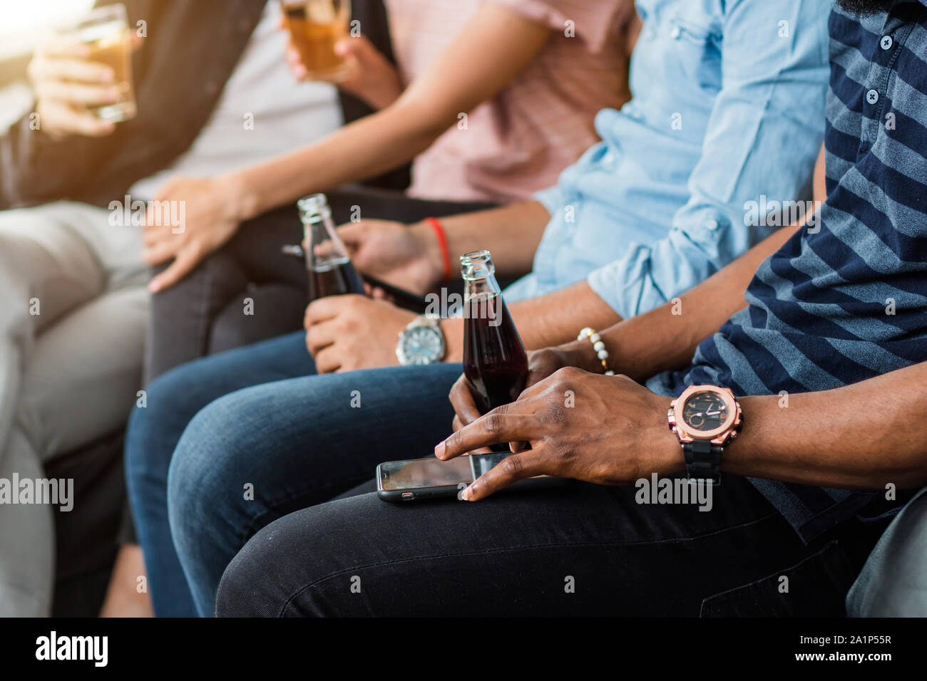 Cropped shot of young african man wearing stylish wrist watch, holding in hand bottle with soft drink and using mobile phone, sitting on sofa with fri Stock Photo