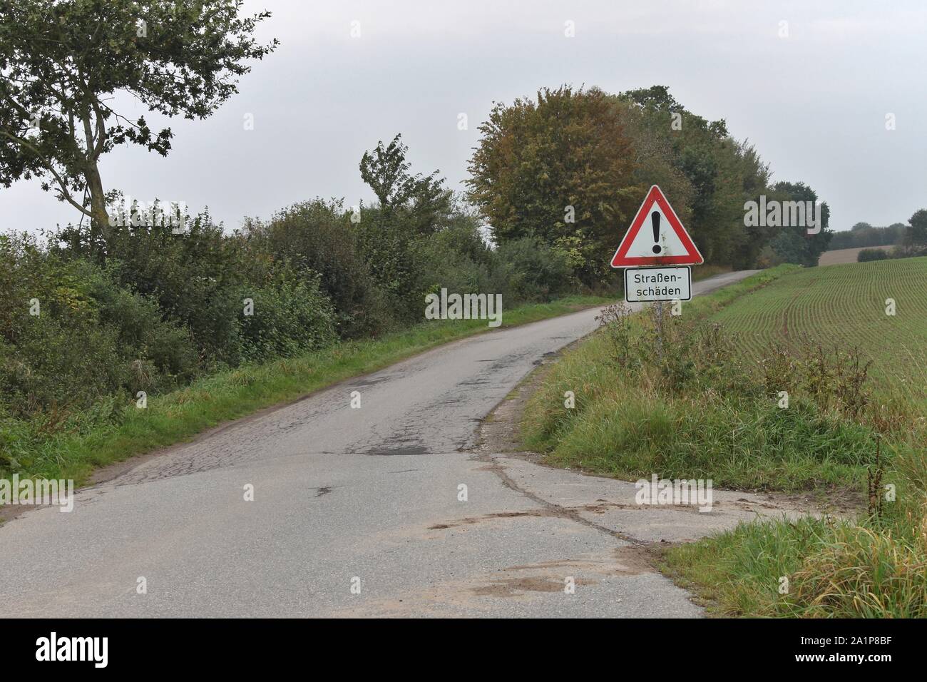 Road sign warning of road surface damage in german. Roadside damage is visible, starting next to the sign. Stock Photo