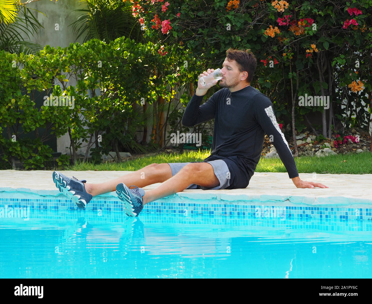 a man after a workout drinks water from a glass while sitting near a swimming pool. Stock Photo