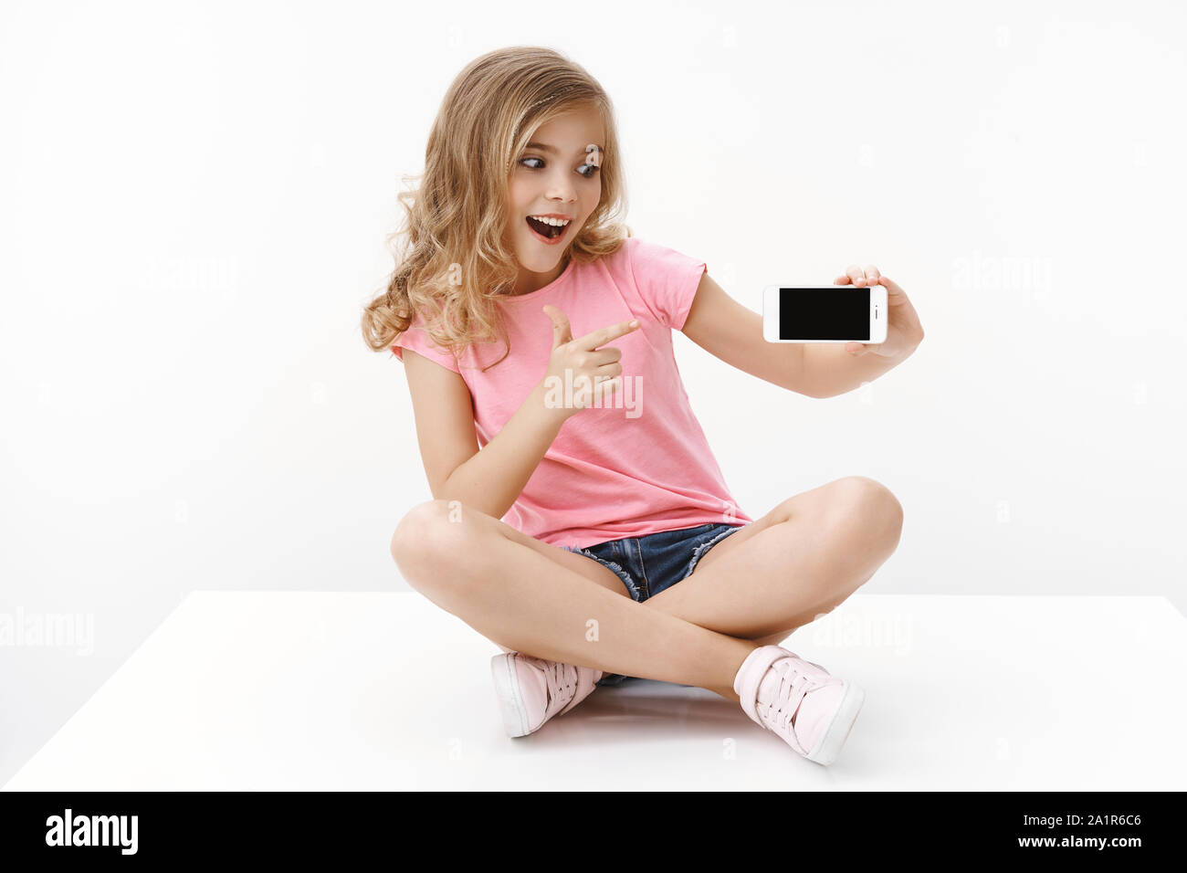 Cheerful excited cute blond little girl sitting on floor with crossed legs, hold smartphone, pointing mobile phone display and looking thrilled Stock Photo