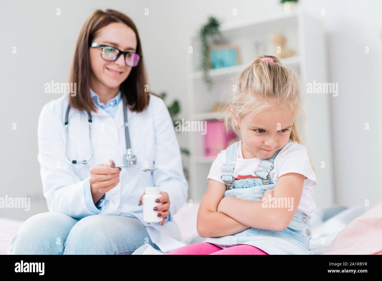 Naughty sick little girl turning away from doctor giving her medicine while both sitting in medical office Stock Photo