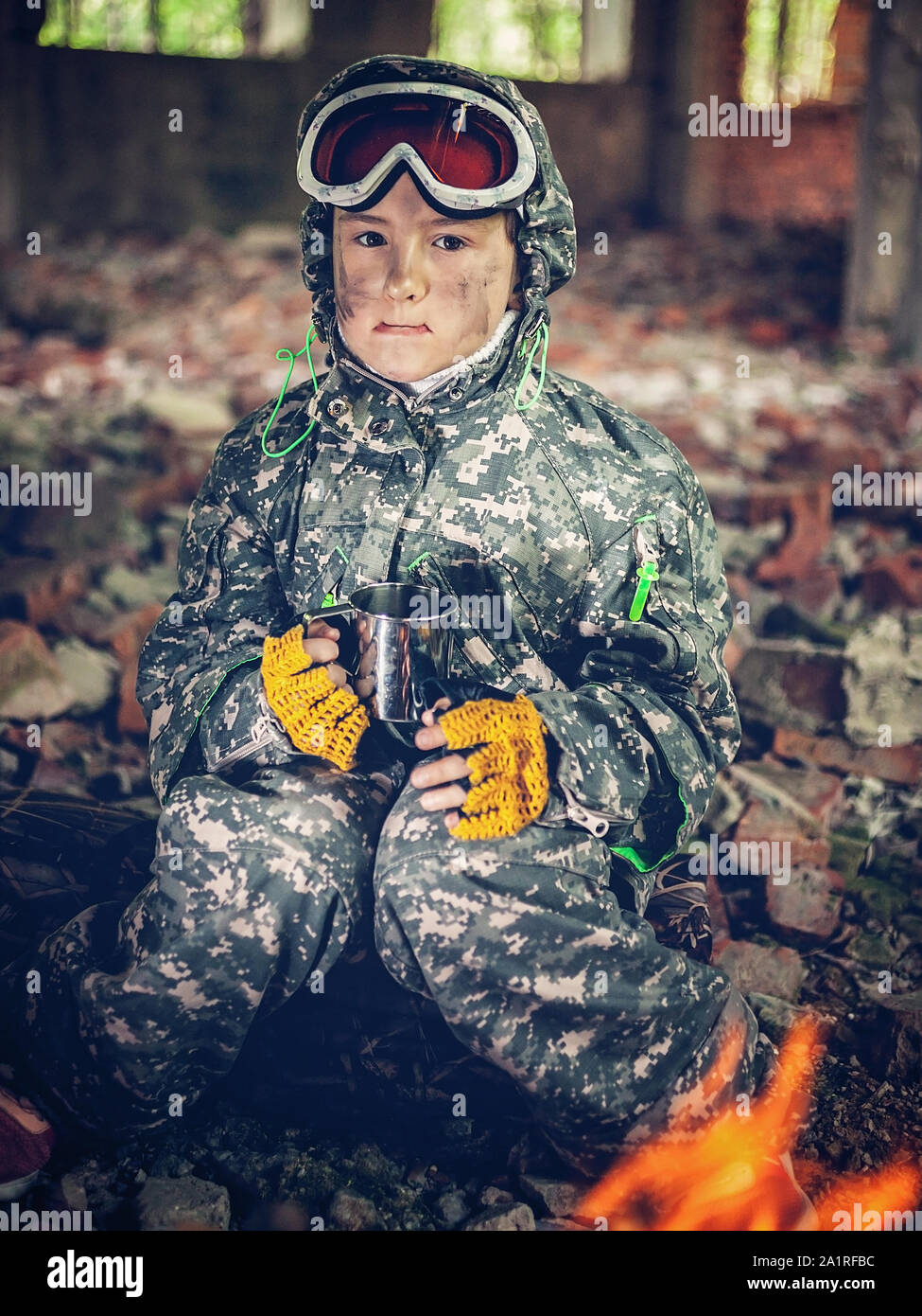 Serious girl in military uniform with mug in her hands sits near a bonfire. Disappointed and upset teenager girl. Disillusioned stalker in abandoned b Stock Photo