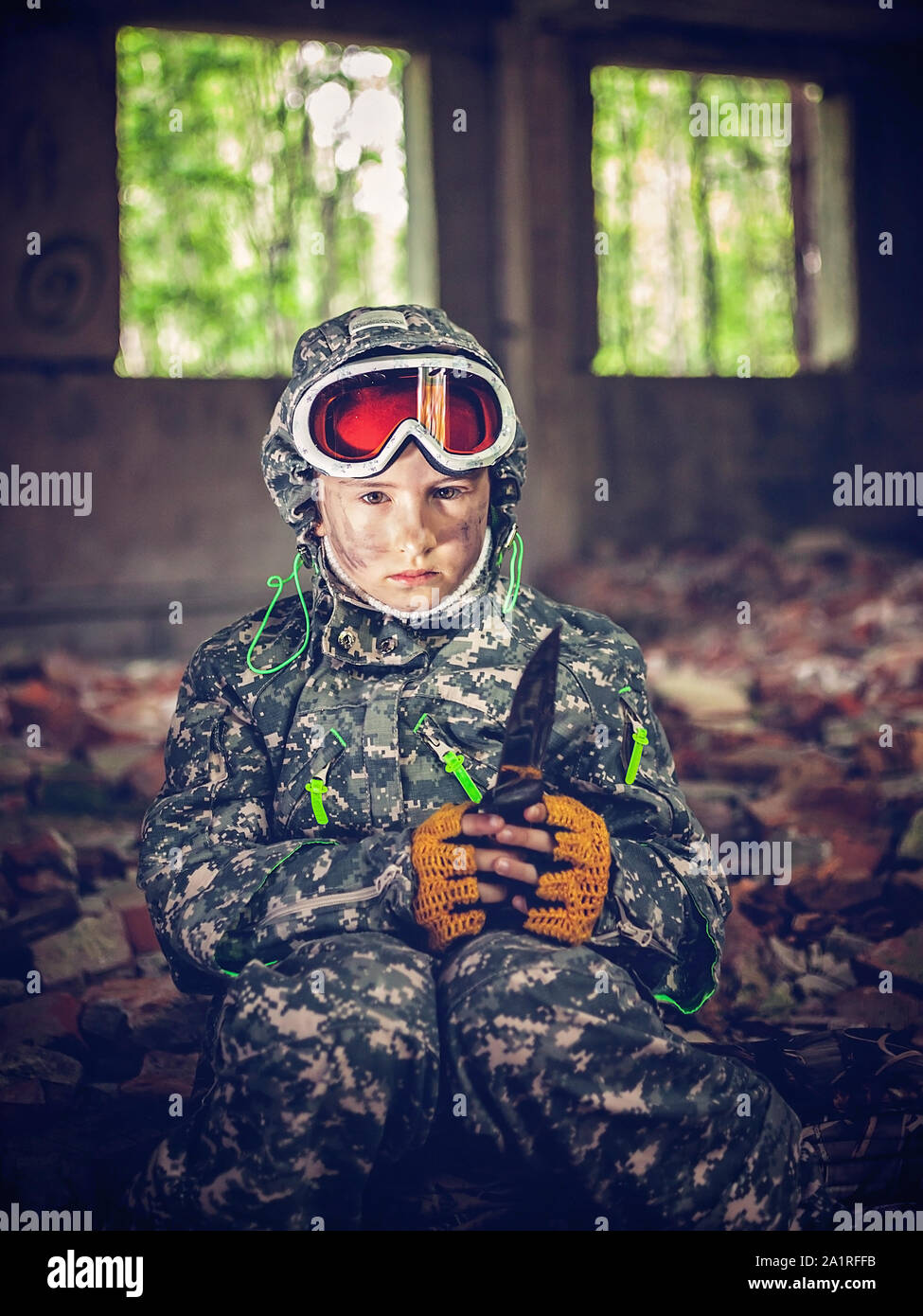 Serious girl in military uniform with knife in her hands sits near a bonfire. Disillusioned stalker in abandoned building. Girl with tired facial expr Stock Photo