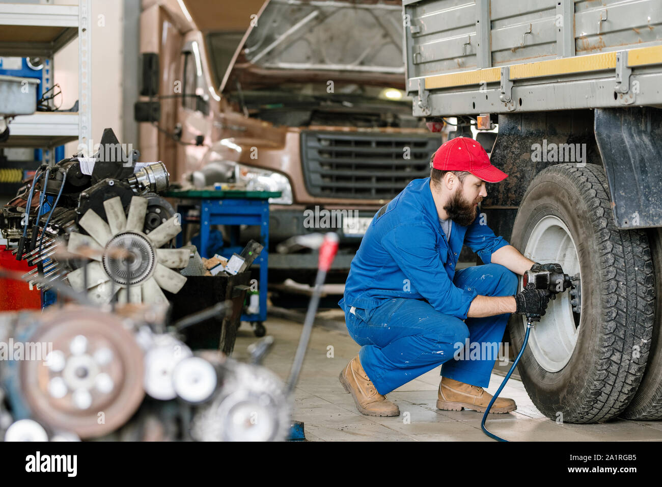 Young technical engineer of car repair service sitting on squats by wheel of large vehicle and drilling Stock Photo