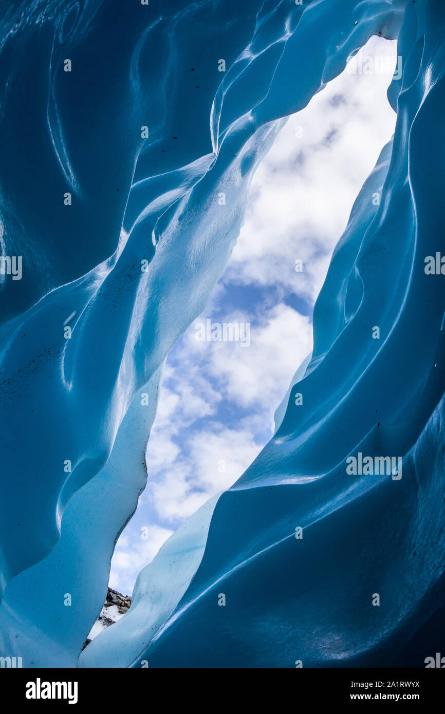 Looking out from a narrow slot canyon ice cave on the Matanuska glacier. Above puffy clouds fill the blue sky. Stock Photo