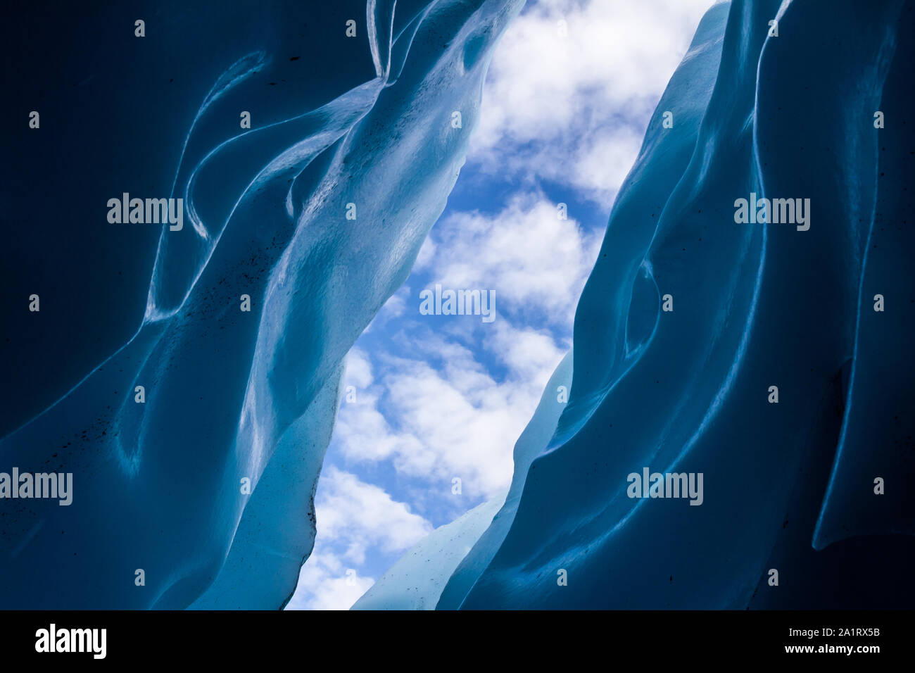 Light bounces off blue ice walls of a crevasse from deep within the Matanuska Glacier in Alaska. The sky above is filled with airy clouds. Stock Photo