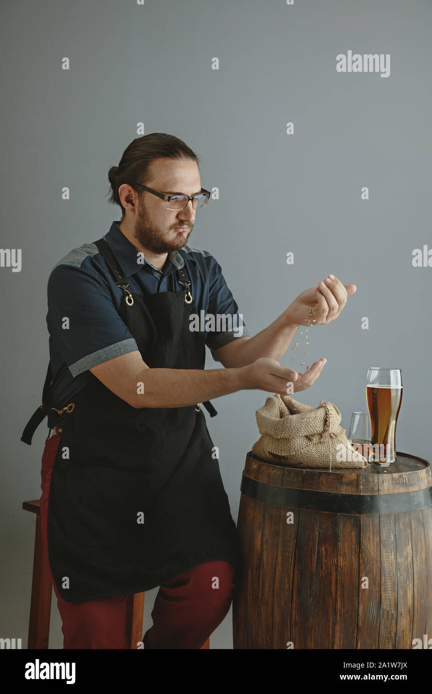Confident young male brewer with self crafted beer in glass on wooden barrel on grey background. Owner of factory man presented his products of brewing. Oktoberfest, drink, alcohol, industry. Stock Photo