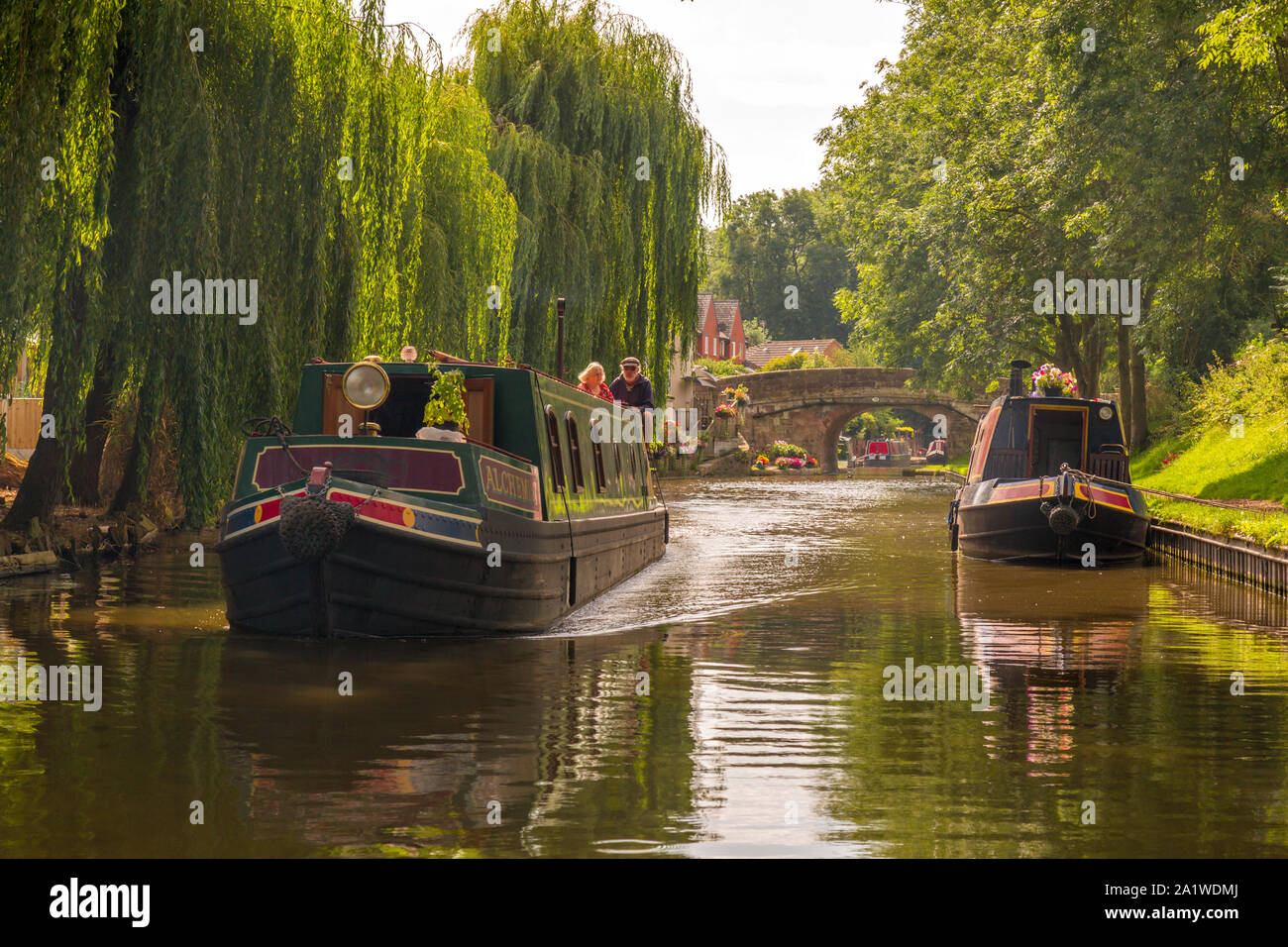 An elderly couple steer a Narrow Boat, or Barge, along The Shropshire Union Canal in England, with another boat moored up. Stock Photo