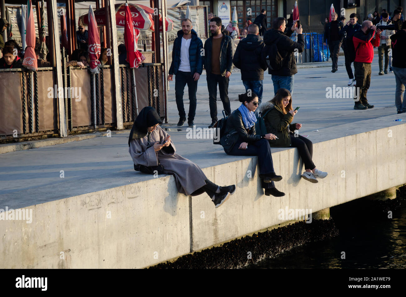 Istanbul, Turkey, March 07,2019: three women sitting and relaxing on stone fence looking at phones, diversity people in crowded place Bosphorus, Stock Photo