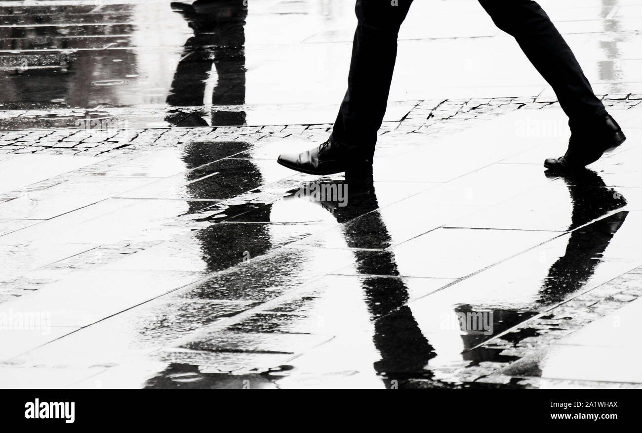 Silhouette and reflection of a man in elegant shoes walking wet city street on s rainy day, legs in black and white Stock Photo