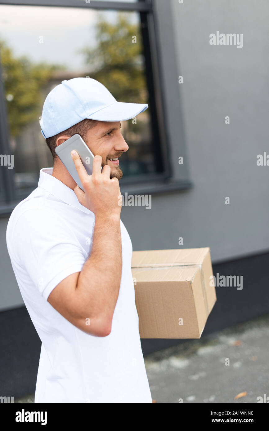 cheerful and bearded delivery man talking on smartphone while holding box Stock Photo