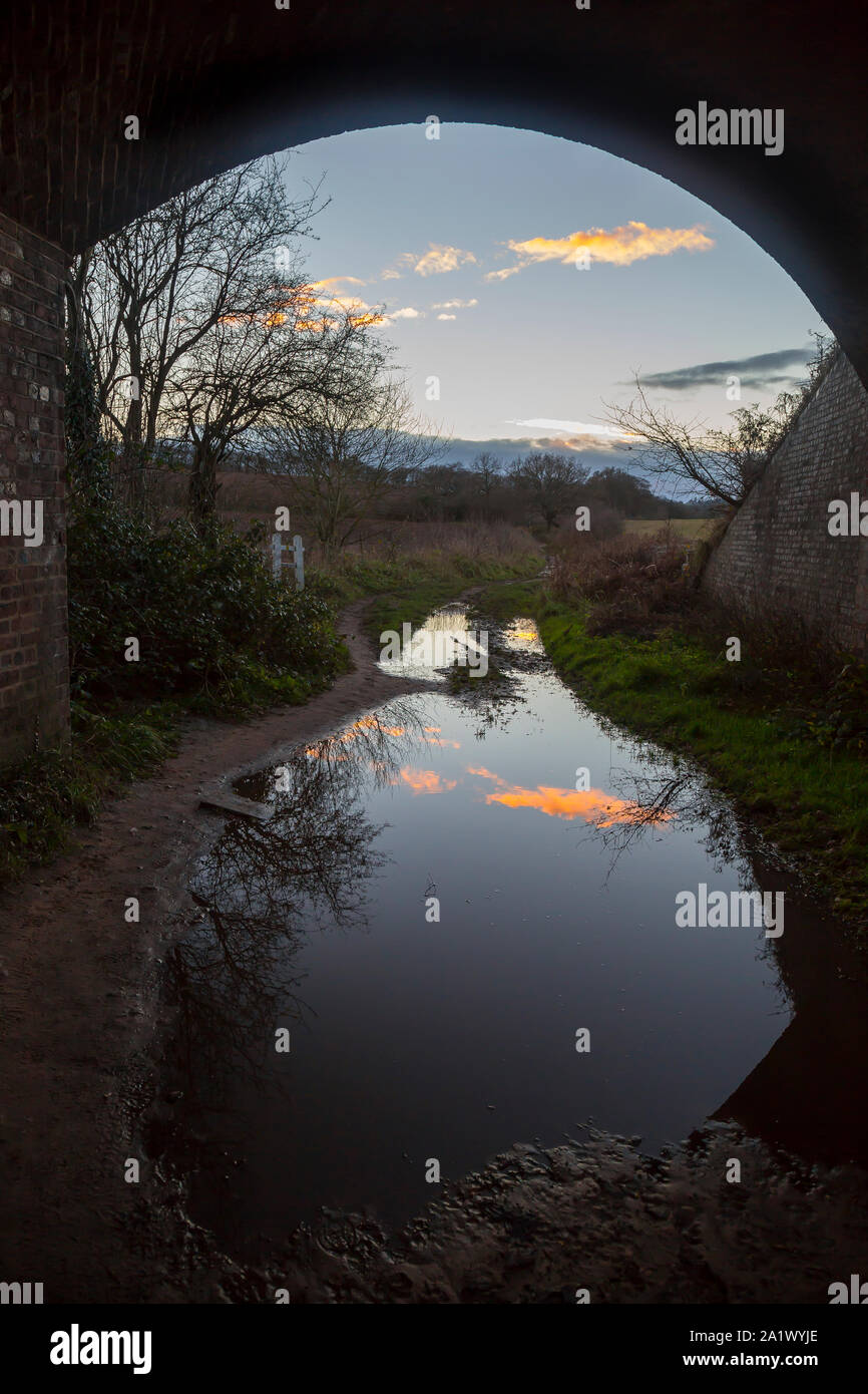 UK countryside on a winter afternoon viewed from under railway arch bridge; blue sky beautifully reflected in a large puddle on ground. Stock Photo