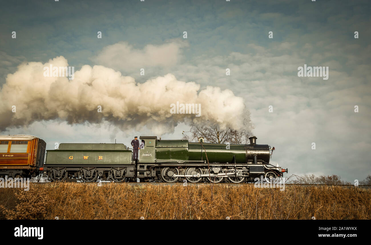 Low angle side view of vintage, UK steam train moving along railway track in a bright winter countryside scene. Steam locomotive puffing. Stock Photo