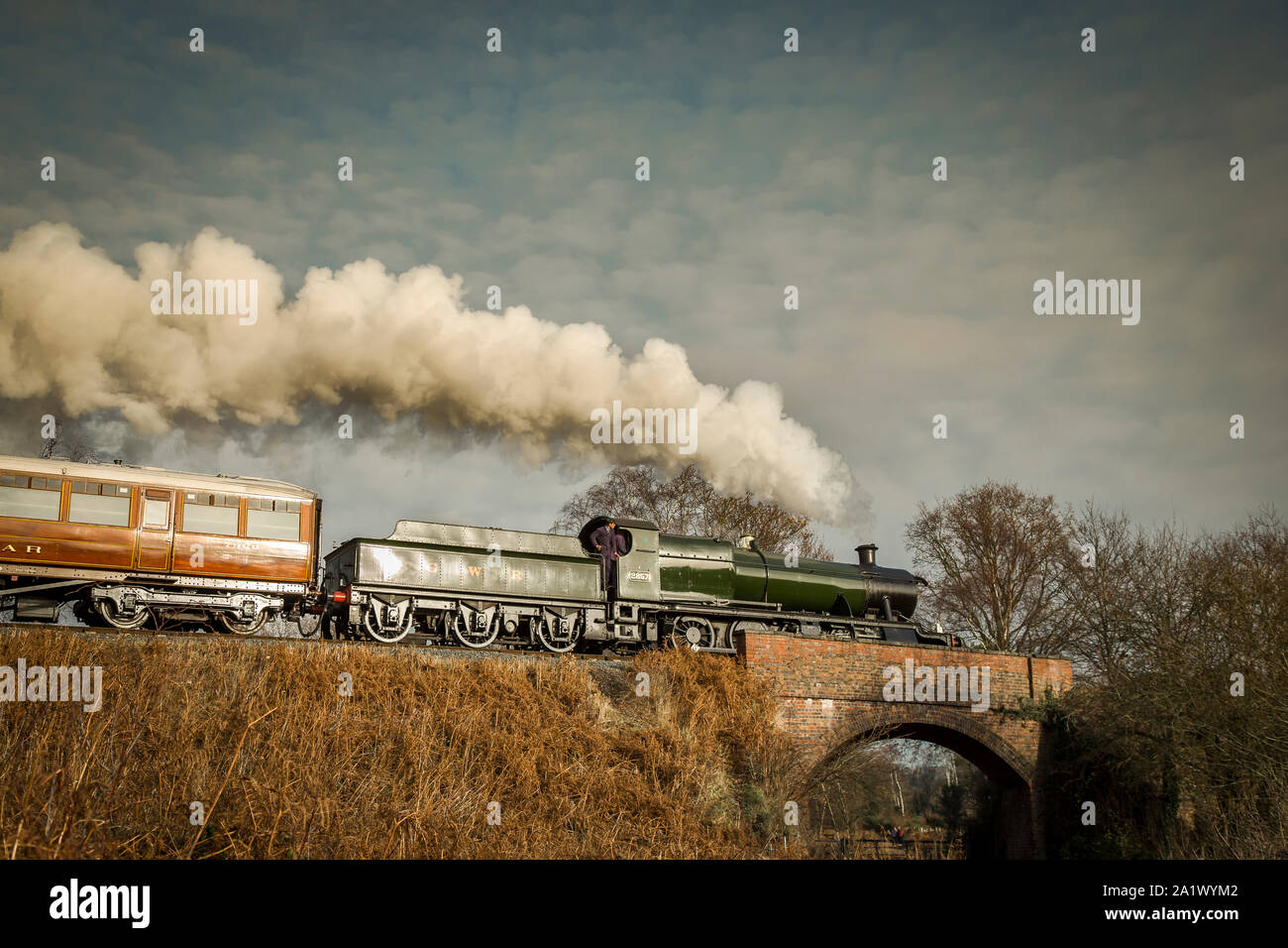 Low-angle side view of vintage, puffing UK steam train moving along the track in a bright, winter countryside scene, steaming over bridge. Stock Photo