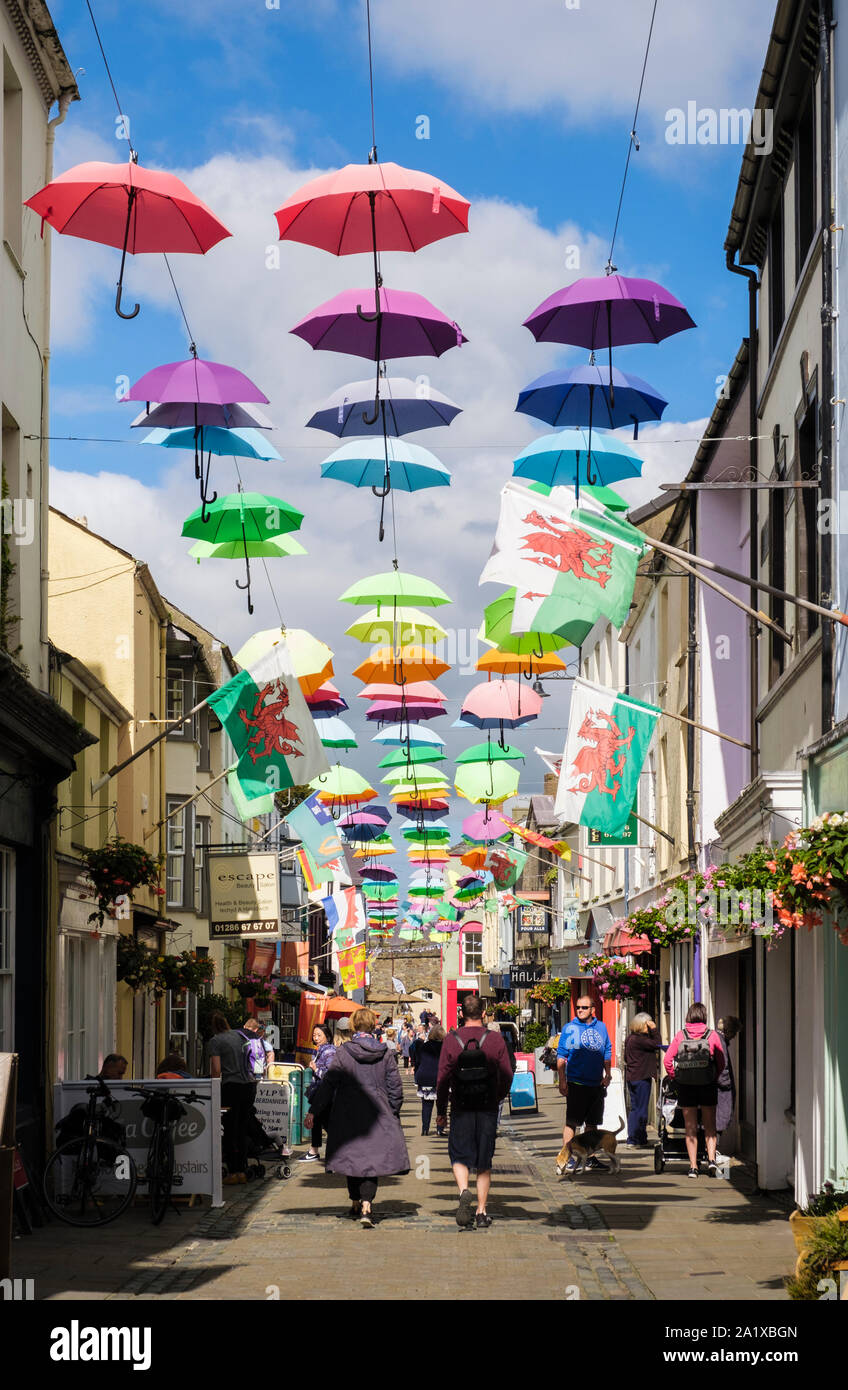 Art installation colourful display of umbrellas hanging above a busy narrow sunlit street in old town. Stryd y Plas, Caernarfon, Gwynedd, Wales, UK Stock Photo