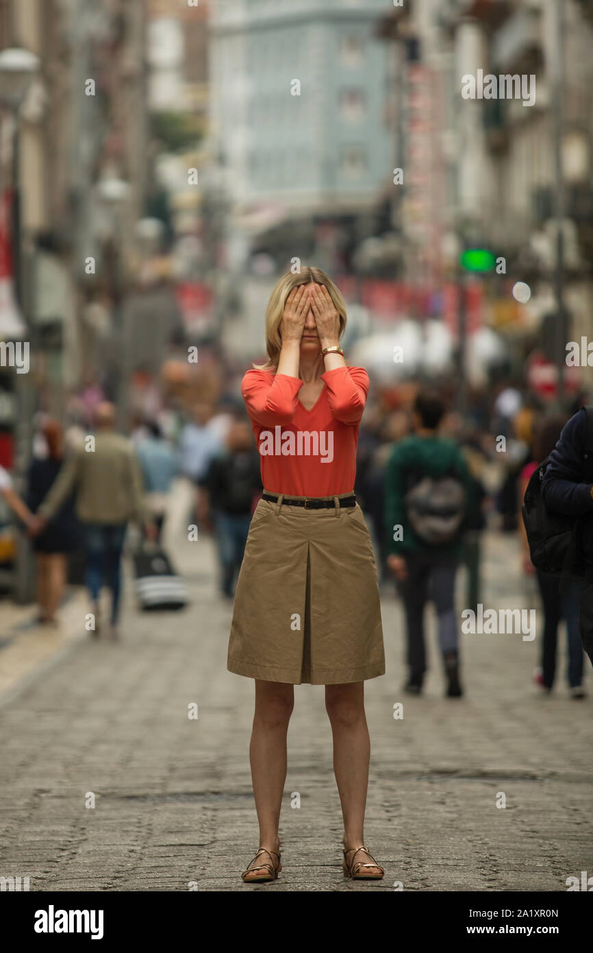 Depressed sad woman covers his eyes with his hands surrounded by people walking in crowded street. Panic attack in public place. Stock Photo