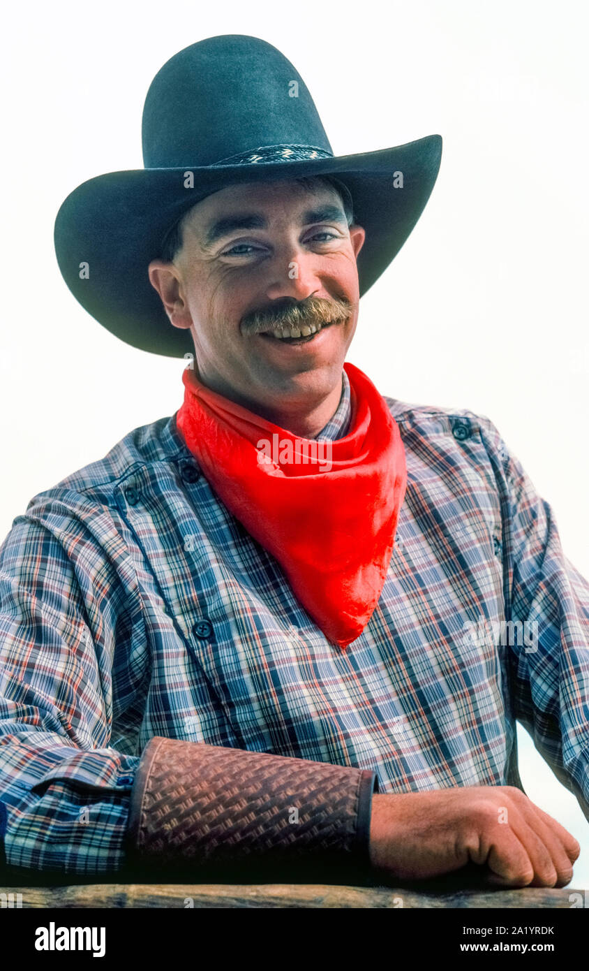 A mustached wrangler smiles while posing for a picture before leading a trail ride on horseback with guests at a dude ranch in the southwestern state of Arizona, USA. His cowboy outfit includes a black ten-gallon hat, a red bandanna around his neck, a long-sleeve plaid shirt, and leather cuffs to protect his wrists and shirtsleeves. The neckerchief is standard attire, and cowboys call it by various names like neck rag, wild rag, buckaroo scarf, and kerchief.  Most common of  its many uses is pulling up the cloth over the face for protection against dust, wind, or inclement weather. Stock Photo
