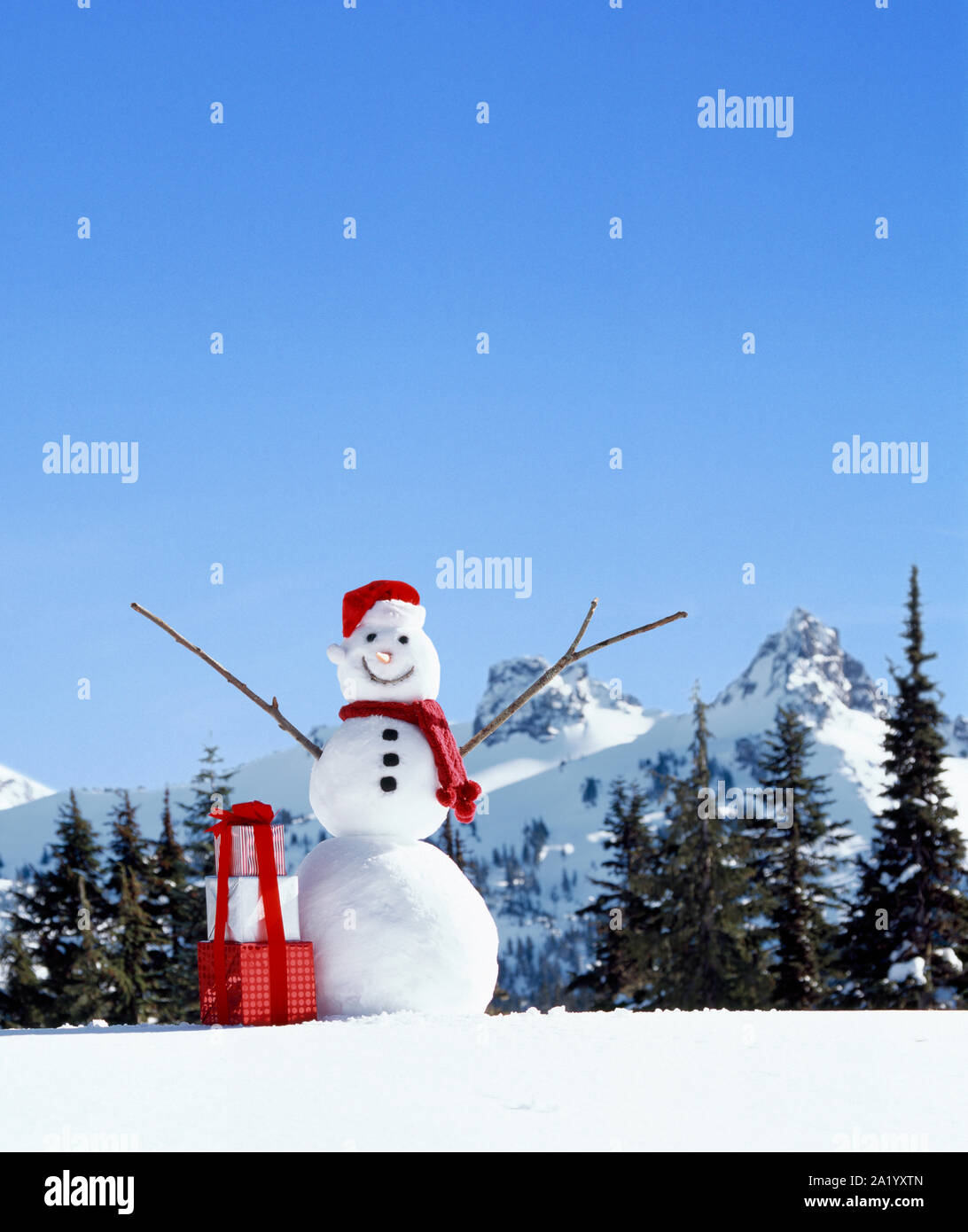 Happy, smiling snowman with Santa hat, stack of festive, wrapped Christmas gifts presents and snowy mountains in background Stock Photo