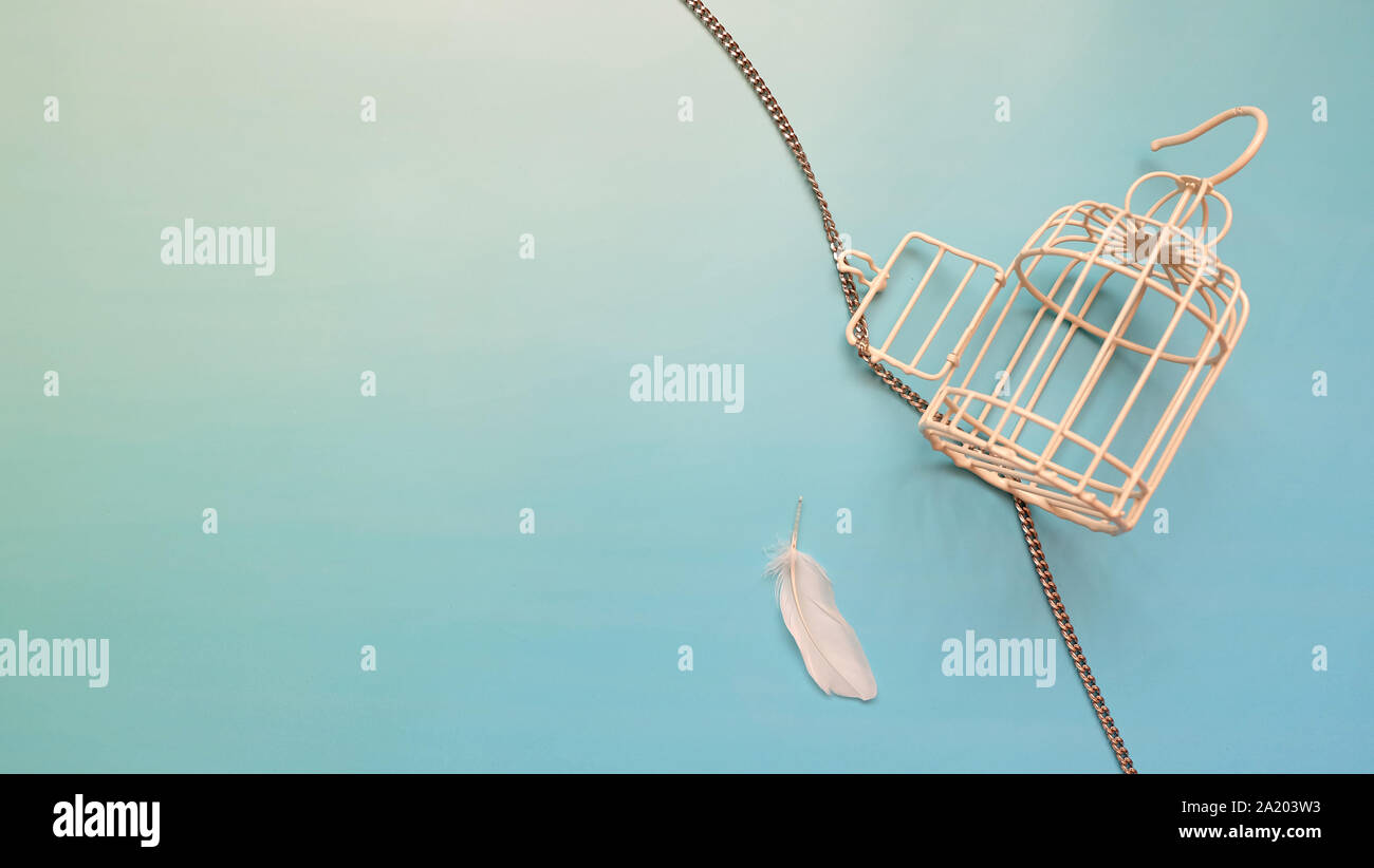 A small empty metal bird cage with its door opened and chain unwind. A single white feather is placed next to the cage. Stock Photo