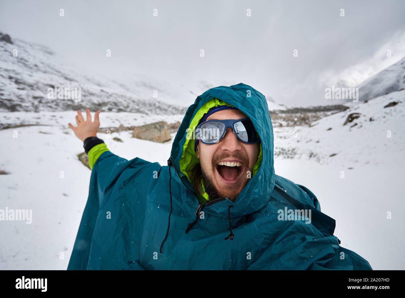 Selfie of smiling hiker in sunglasses and blue raincoat poncho in snowy mountains. Outdoor travel concept Stock Photo