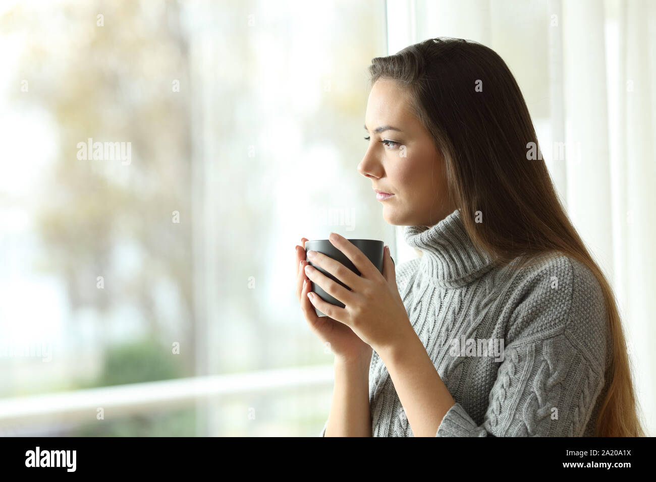 Worried woman holding a coffee mug looks through a window at home in winter Stock Photo