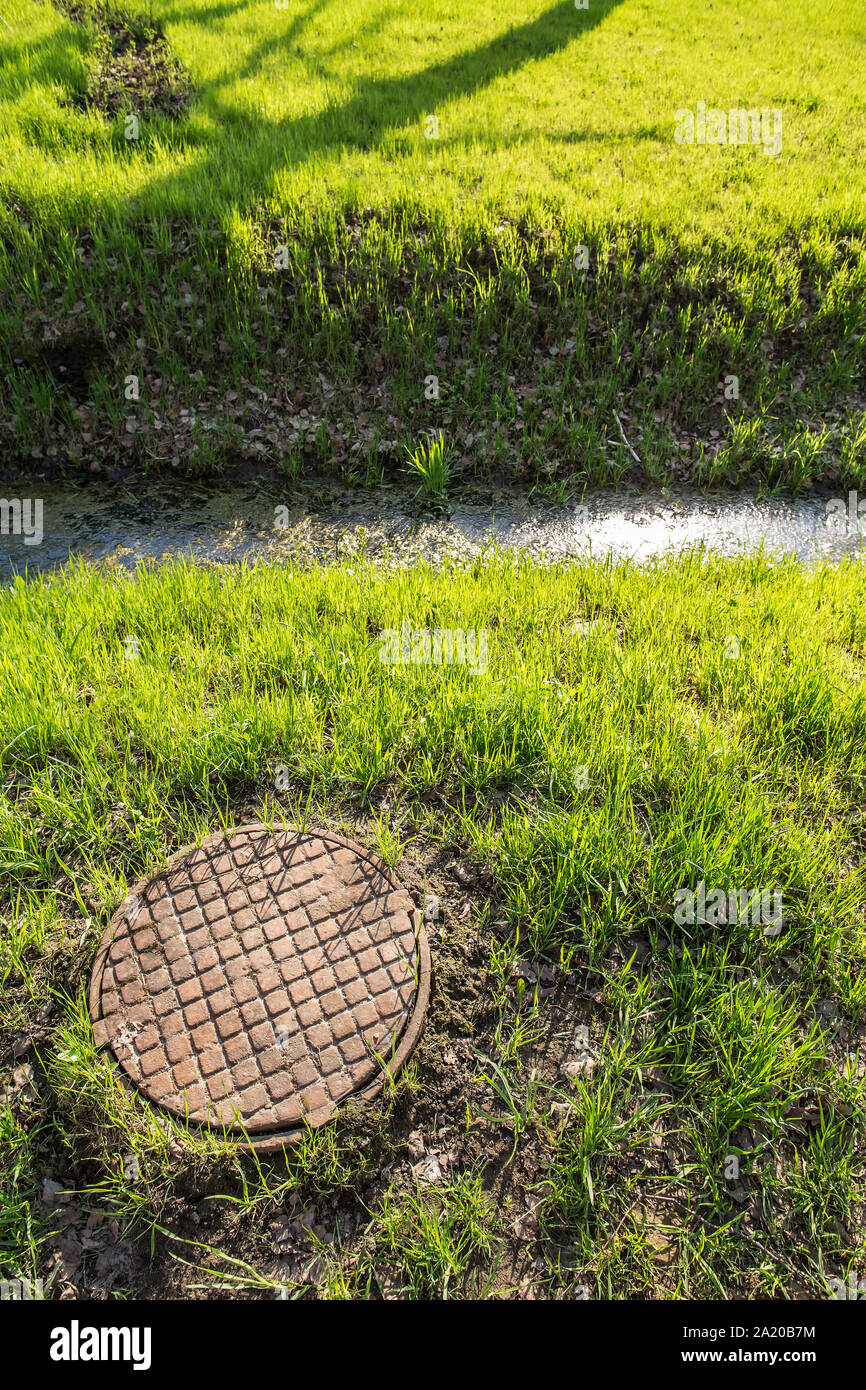 old rusty the sewer hatch with cover and a gutter with water and ooze is in the park among fresh spring grass Stock Photo
