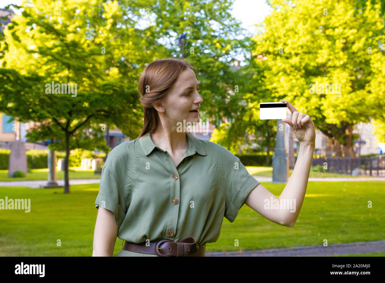 Happy young blonde girl in a green dress with a credit card on a park ...