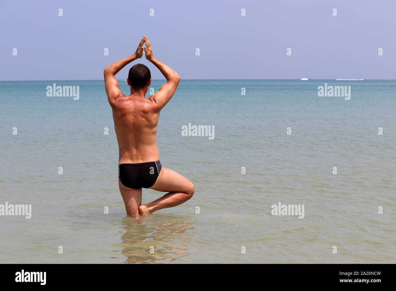Yoga on the beach, man stands in a tree pose in the blue water of tropical sea. Balance with nature, fitness, relaxation and exercise at vacation Stock Photo