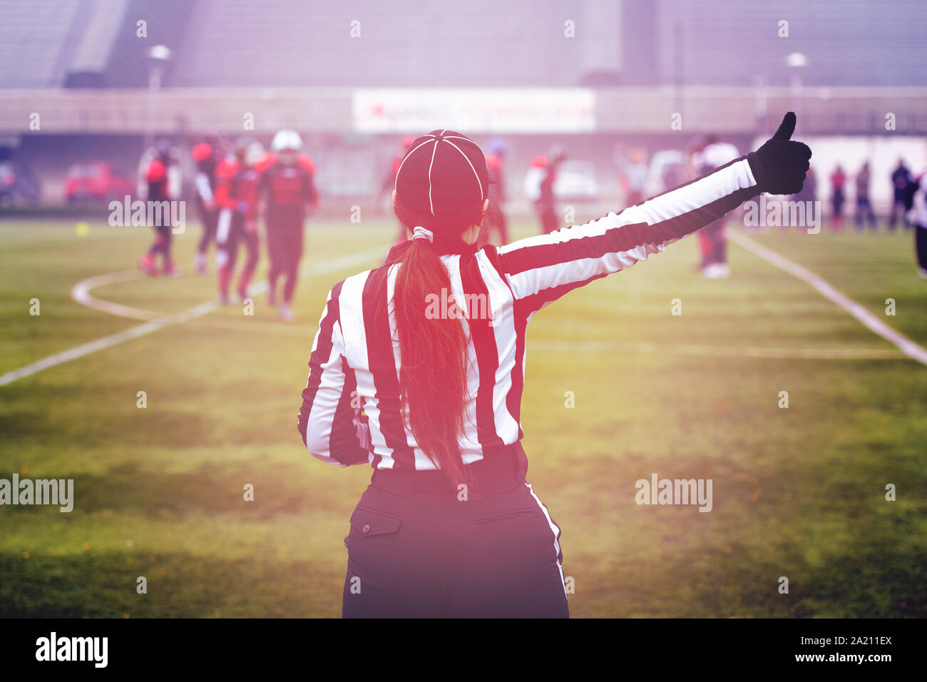 rear view of female american football referee giving signals to professional players during match on the stadium field Stock Photo