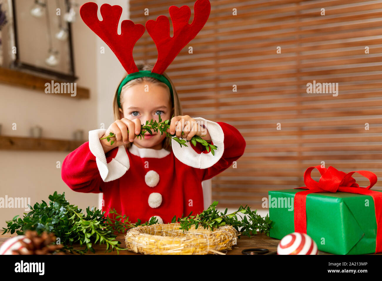 Cute preschooler girl dressed up for xmas making christmas wreath in living room. Christmas decoration family fun concept. Stock Photo