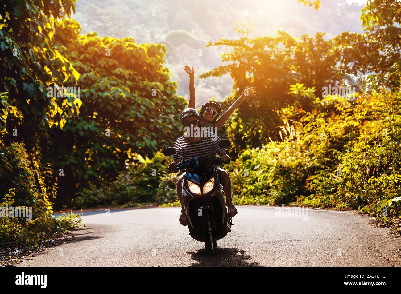 Happy couple rides on motorbike or scooter against sunset tropic road Stock Photo