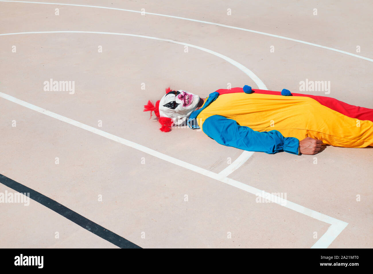 a scary clown, wearing a colorful yellow, red and blue costume, lying face up on an outdoor basketball court Stock Photo