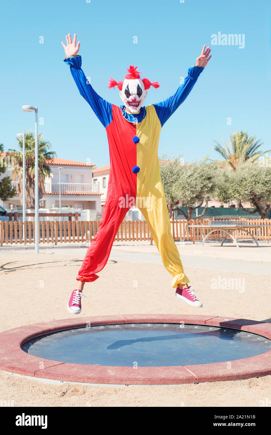 a scary clown wearing a colorful yellow, red and blue costume bouncing on a trampoline in an outdoor public playground Stock Photo