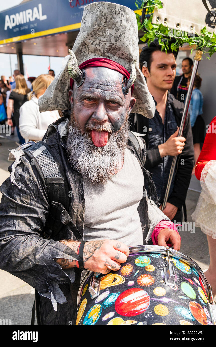 Mature man, drummer, with horns attached to side of head, dressed as a pirate looking directly at viewer and poking tongue out. Pirate Day, Hastings. Stock Photo