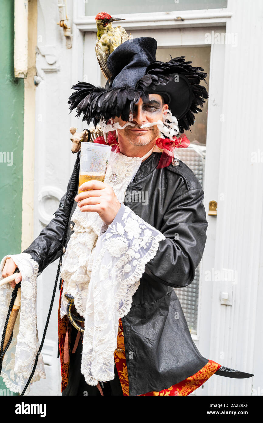 Pirate Day in Hastings, UK. Smiling man dressed as pirate with stuffed wood pecker on hat, raising a glass of beer to viewer as he poses. Eye-contact. Stock Photo