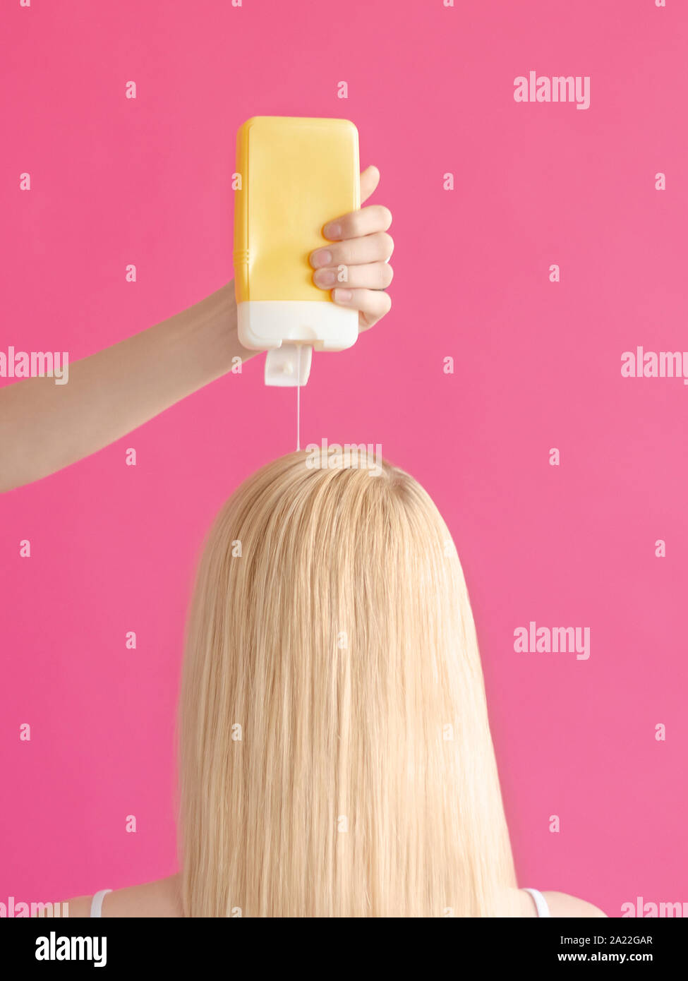 Washing hair  Woman's hand is pouring shampoo from yellow bottle on the head of a long-haired blonde girl against pink background Stock Photo