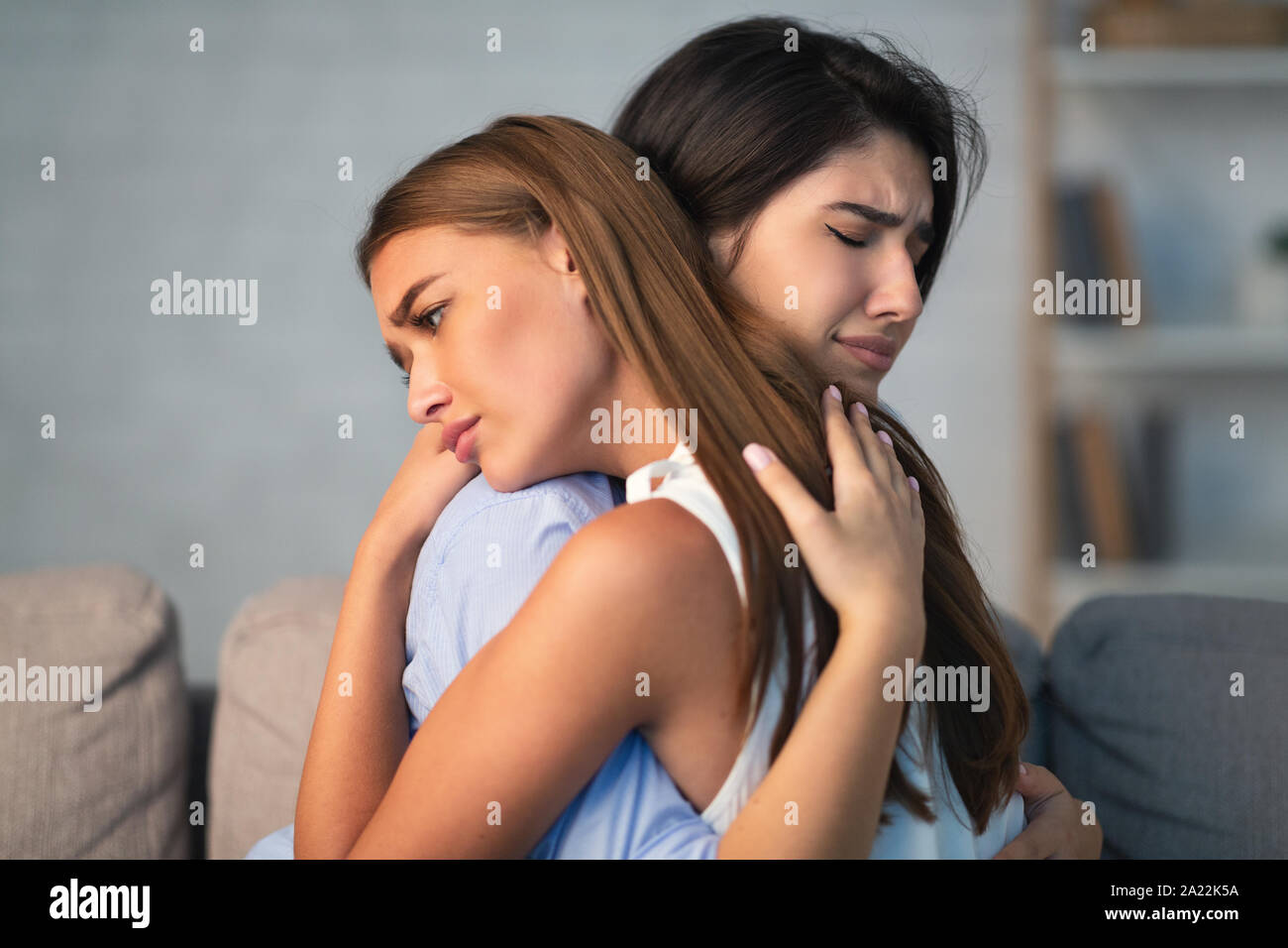 Compassionate Girl Hugging Crying Friend Supporting Her After Breakup With Boyfriend Sitting On Sofa Indoor. Selective Focus Stock Photo