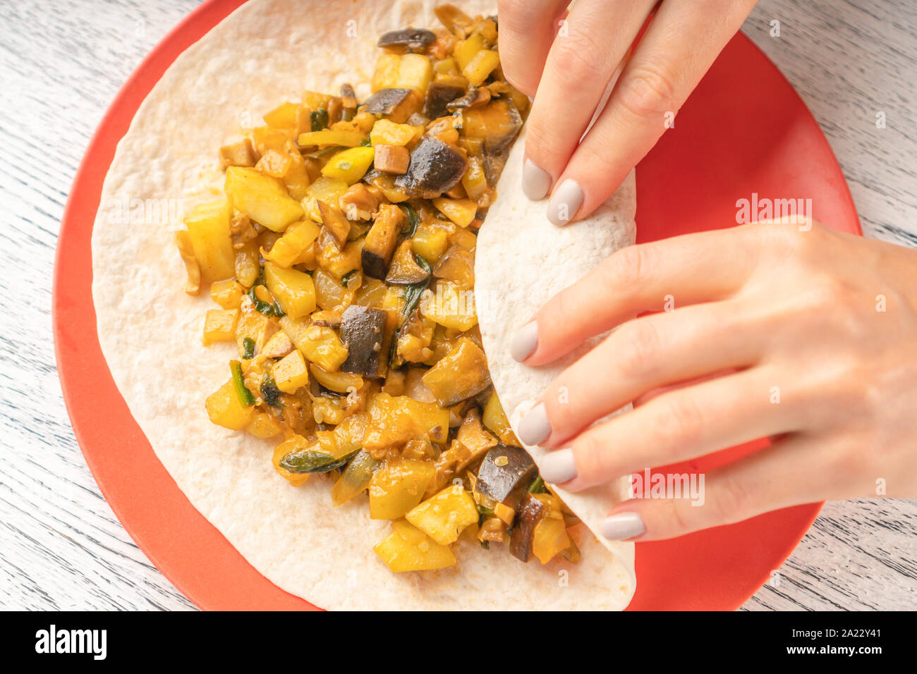 Girl prepares food from pita bread and vegetable puree with chopped eggplant and zucchini on a red plate on a black-white wooden table. Cooking nation Stock Photo