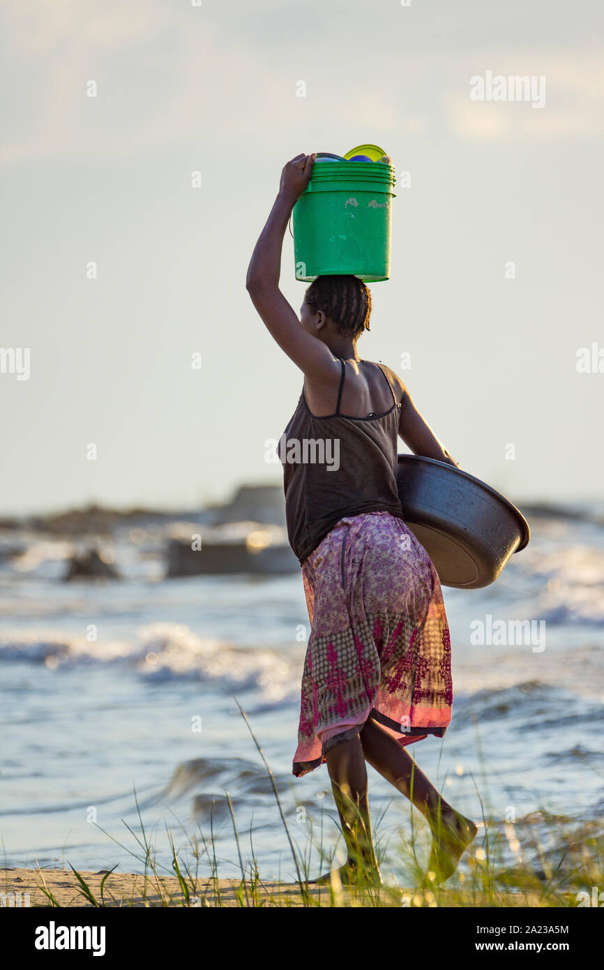 Woman walking on the shore of Lake Malawi carrying a plastic basket and metal washing bowl. Stock Photo