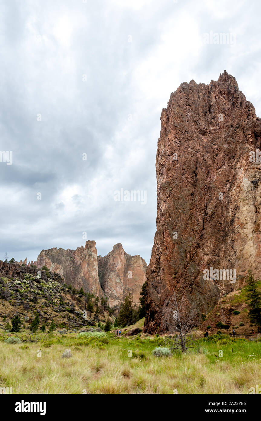 Smith Rock State Park with high cliffs of basalt and tuff, ideal for rock climbing. Vertical shot with moody sky, green grass below. Stock Photo