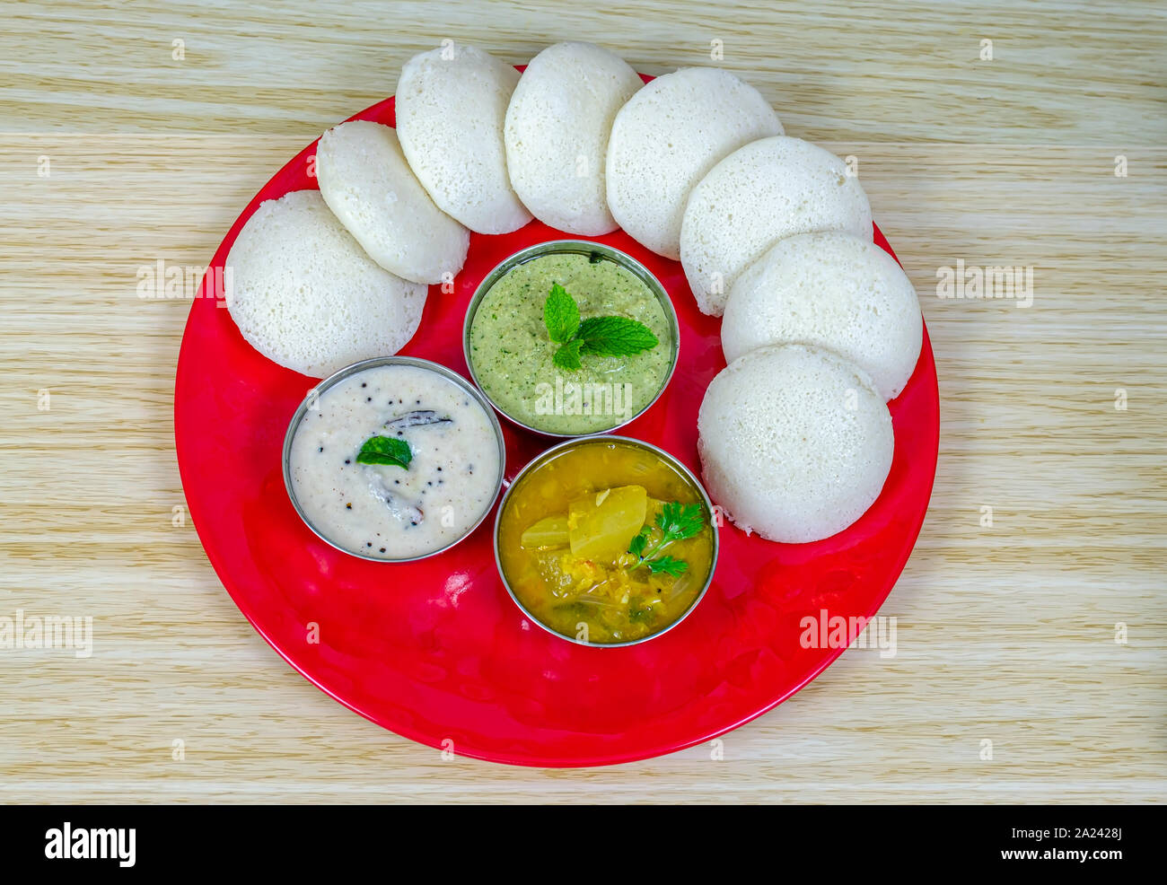 Idlis arranged in a circular formation with white and green chutneys, and sambar in a red plate. Stock Photo