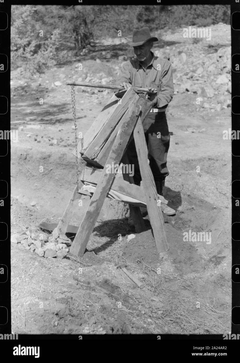 Prospector operating dry washer called a papago. This dry washer is the old Indian method of washing for gold. The rocks and dirt are placed in the higher compartment. Below and to the back is a bellows made of canvas. This is pumped to blow through the screening and thus blow away loose dirt while the heavy gold remains. Pinos Altos, New Mexico. Stock Photo