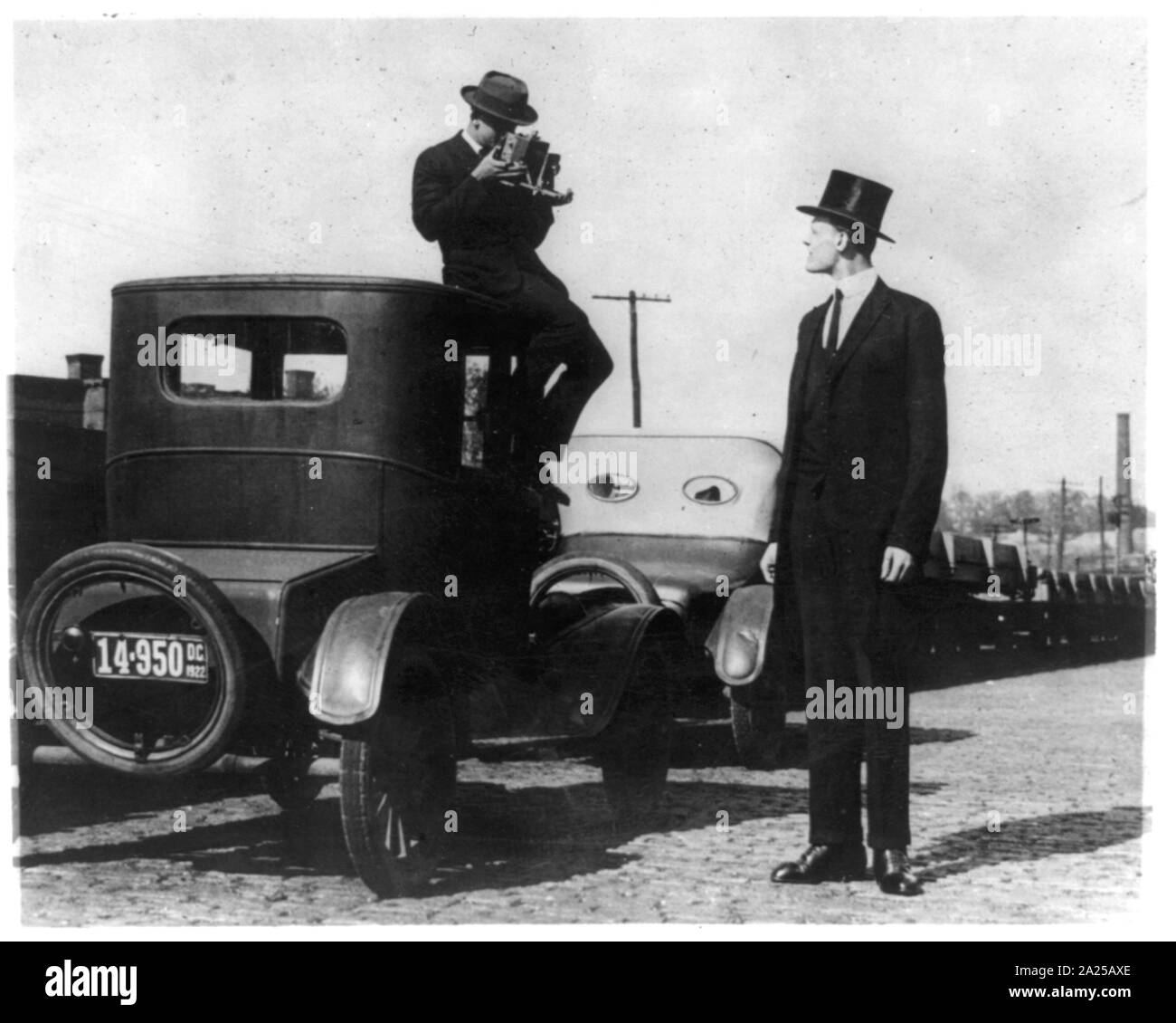 Photographer seated on top of car photographing tall man in top hat Stock Photo