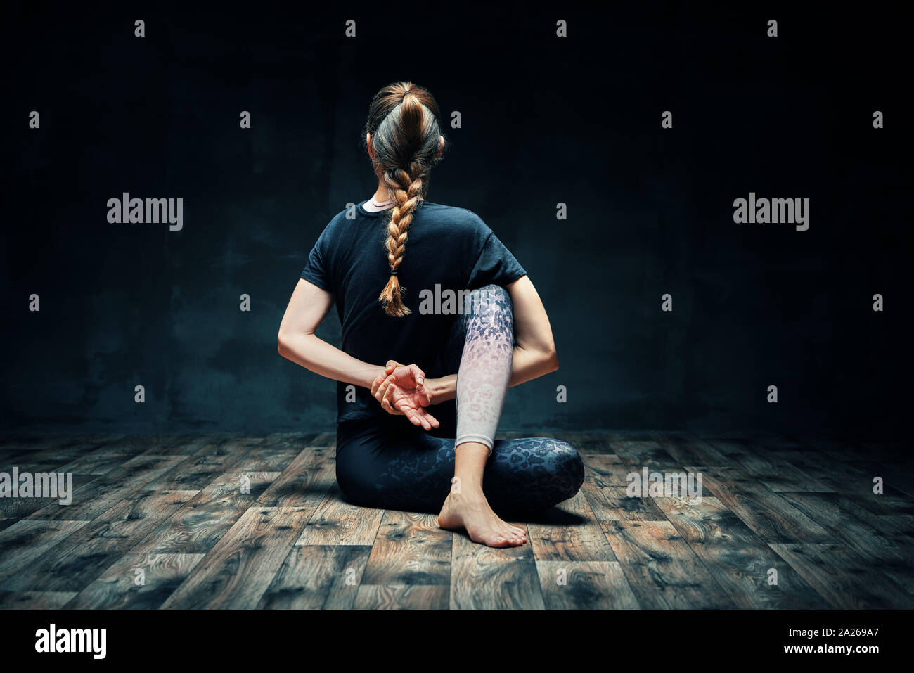 Back view of young woman doing yoga asana half lord of the fishes pose on dark room. Practicing yoga, wellbeing and healthy lifestyle. Ardha Matsyendr Stock Photo