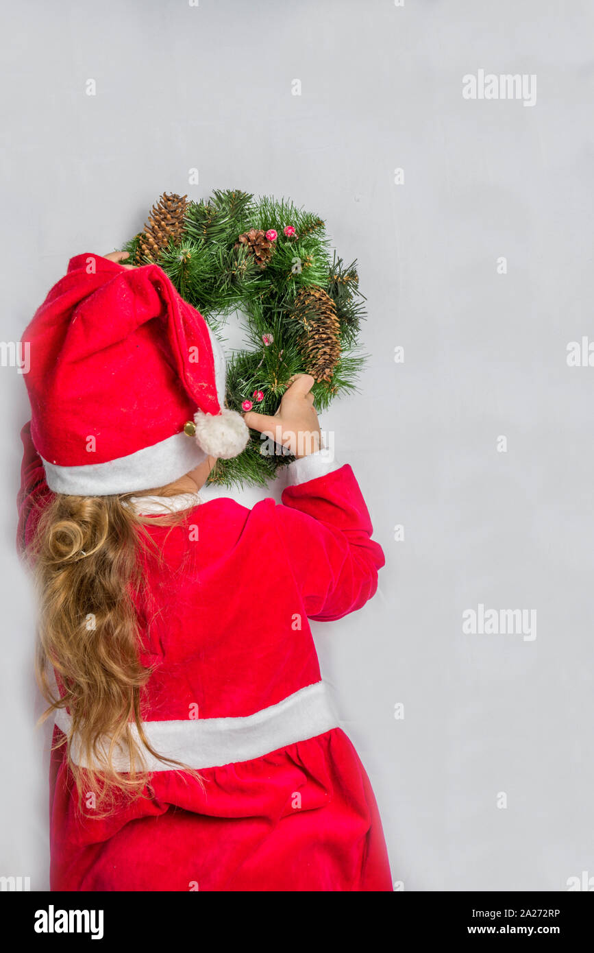 Little blonde cute girl in santa dress hangs a Christmas wreath on wall ...